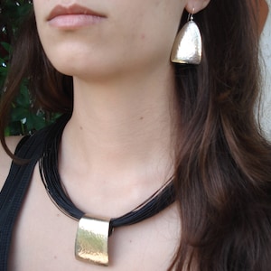 May include: A woman wearing a black top with a layered black cord necklace and a gold pendant. She is also wearing silver earrings with a hammered texture.