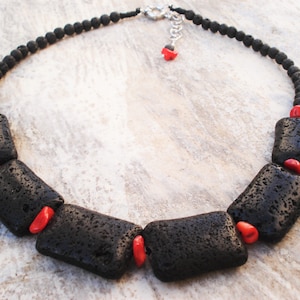 Puede incluir: Un collar de piedra de lava negra con cuentas de coral rojo. El collar está hecho de pequeñas cuentas redondas de piedra de lava y cuentas rectangulares de piedra de lava más grandes. Las cuentas de coral están intercaladas entre las cuentas de piedra de lava.