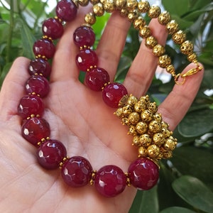 Puede incluir: Un collar de piedras preciosas color burdeos con cuentas doradas y un colgante dorado. El collar está hecho de cuentas de piedras preciosas facetadas y tiene un colgante dorado texturizado.