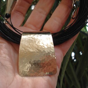 May include: A long, black leather necklace with a hammered silver pendant. The pendant is rectangular and has a textured surface.