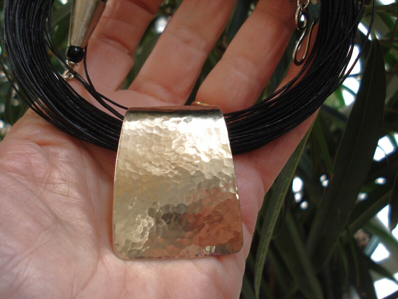 May include: A long, black leather necklace with a hammered silver pendant. The pendant is rectangular and has a textured surface.