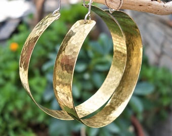 Puede incluir: Un par de pendientes de aro grandes y circulares hechos de metal pulido en tono dorado. La superficie de cada aro presenta una textura martillada distintiva, creando una apariencia reflectante y facetada. Los pendientes cuelgan de ganchos plateados.