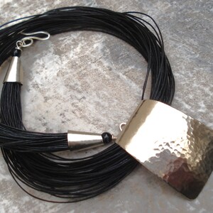 May include: A long black cord necklace with a hammered silver pendant. The pendant is rectangular and has a textured surface. The necklace has a silver clasp and a tassel at the end.