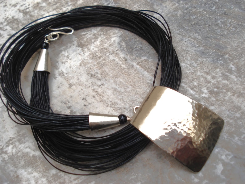 May include: A long black cord necklace with a hammered silver pendant. The pendant is rectangular and has a textured surface. The necklace has a silver clasp and a tassel at the end.