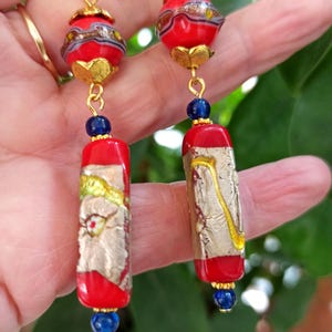 May include: A pair of dangle earrings with red and gold glass beads and blue glass accents. The beads are decorated with gold leaf and have a textured surface.