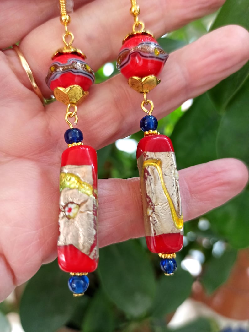 May include: A pair of dangle earrings with red and gold glass beads and blue glass accents. The beads are decorated with gold leaf and have a textured surface.