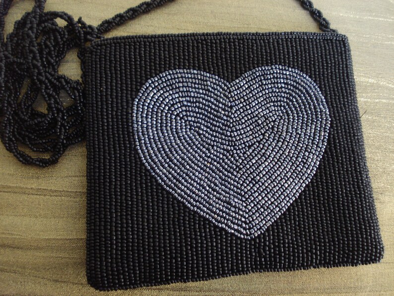 Puede incluir: Un bolso de noche negro de cuentas con un dise&ntilde;o de coraz&oacute;n plateado. El bolso tiene una larga correa negra de cuentas.