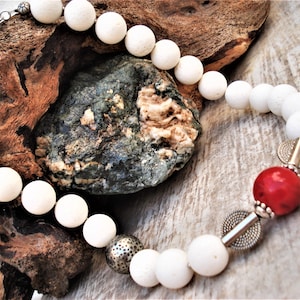 May include: White bead necklace with a red coral bead and silver accents. The necklace is made of natural materials and has a rustic, bohemian style.