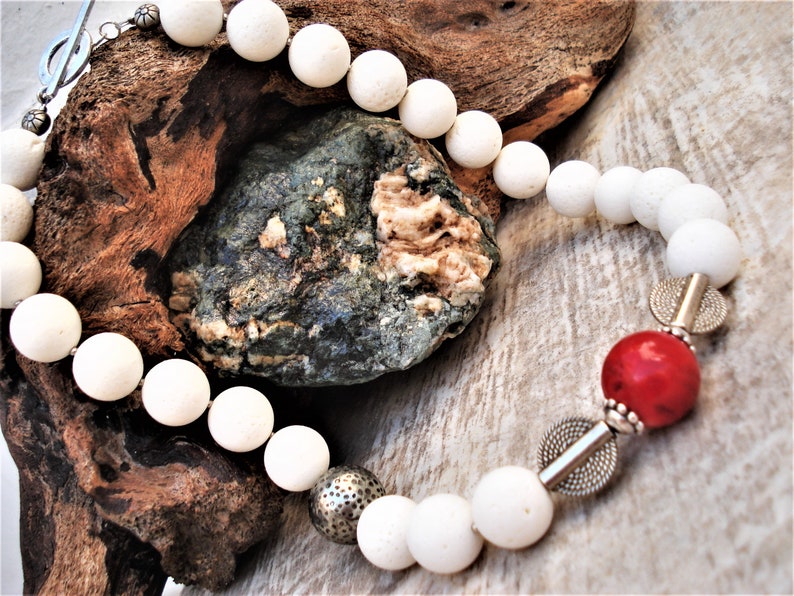 May include: White bead necklace with a red coral bead and silver accents. The necklace is made of natural materials and has a rustic, bohemian style.