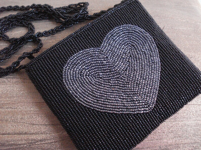 Puede incluir: Un bolso de noche negro de cuentas con un dise&ntilde;o de coraz&oacute;n plateado. El bolso tiene una correa de cadena.