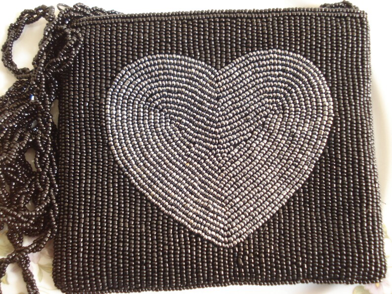 Puede incluir: Un bolso de noche negro de cuentas con un dise&ntilde;o de coraz&oacute;n plateado.
