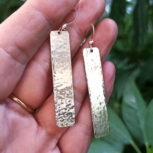 Puede incluir: Un par de pendientes de metal plateado martillado. Cada pendiente tiene forma rectangular alargada con una superficie texturizada y un pequeño gancho para la oreja. Los pendientes se muestran sobre un fondo verde borroso.