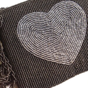 Puede incluir: Un bolso de mano negro de cuentas con un dise&ntilde;o de coraz&oacute;n plateado. El bolso tiene un detalle de flecos.