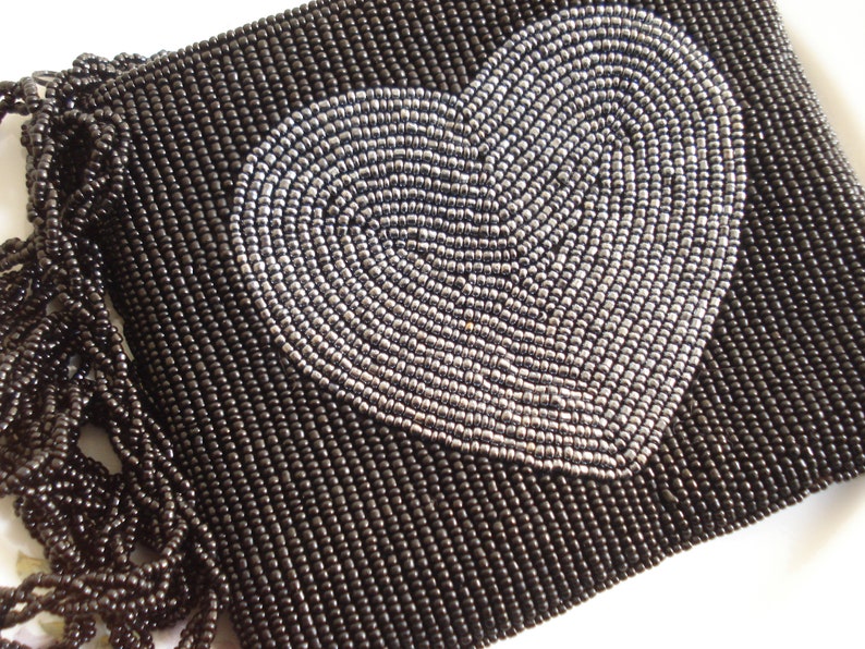 Puede incluir: Un bolso de mano negro de cuentas con un dise&ntilde;o de coraz&oacute;n plateado. El bolso tiene un detalle de flecos.