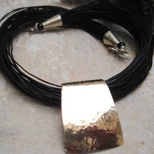 May include: A black leather cord necklace with a hammered gold pendant. The necklace is made of multiple strands of leather cord and has a simple, elegant design.