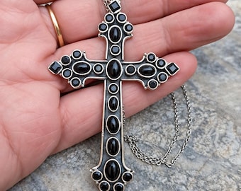 Op de afbeelding: Een grote zilveren kruishanger aan een bijpassende zilveren ketting, voorzien van meerdere zwarte ovale en ronde cabochonstenen in een metalen frame, met kleine vierkante zwarte stenen aan de uiteinden van het kruis.