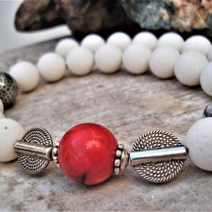 May include: A white beaded necklace with a red coral bead and silver accents. The necklace is made of natural materials and has a rustic, bohemian style.