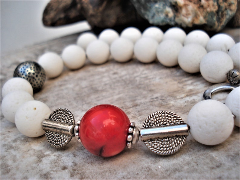 May include: A white beaded necklace with a red coral bead and silver accents. The necklace is made of natural materials and has a rustic, bohemian style.