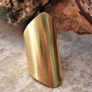 Puede incluir: Un anillo ancho de metal de color dorado con un acabado liso y cepillado. El anillo está ligeramente curvado y tiene un borde redondeado.