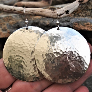 Puede incluir: Un par de pendientes circulares grandes de metal plateado con una textura martillada y con hoyuelos en toda la superficie. Cada pendiente cuelga de un gancho de alambre fino y curvado de color plateado en el borde superior.