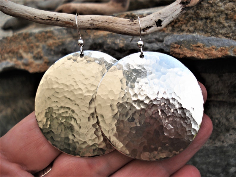Puede incluir: Un par de pendientes circulares grandes de metal plateado con una textura martillada y con hoyuelos en toda la superficie. Cada pendiente cuelga de un gancho de alambre fino y curvado de color plateado en el borde superior.