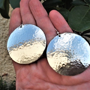 Puede incluir: Un par de pendientes circulares grandes de color plateado con una textura de superficie martillada y reflectante. Cada pendiente cuelga de una peque&ntilde;a cuenta de metal y un gancho, creando un aspecto met&aacute;lico muy pulido.