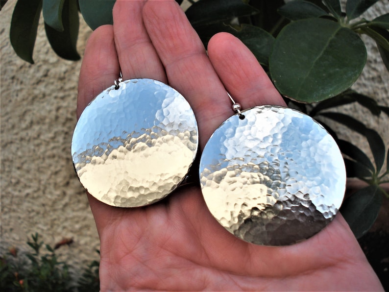 Puede incluir: Un par de pendientes circulares grandes de color plateado con una textura de superficie martillada y reflectante. Cada pendiente cuelga de una peque&ntilde;a cuenta de metal y un gancho, creando un aspecto met&aacute;lico muy pulido.