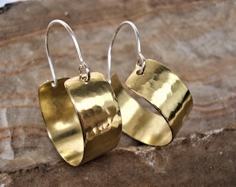 Puede incluir: Un par de pendientes de aro con bandas anchas y curvas de metal dorado martillado. Cada pendiente cuelga de un fino gancho de alambre plateado que pasa por un pequeño orificio en la parte superior de la banda metálica.