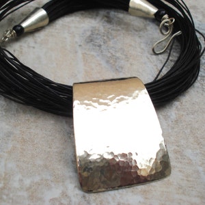 May include: A black leather cord necklace with a large, hammered silver pendant. The pendant is rectangular and has a textured surface.