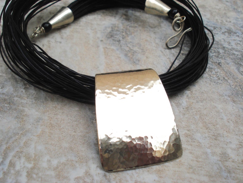 May include: A black leather cord necklace with a large, hammered silver pendant. The pendant is rectangular and has a textured surface.
