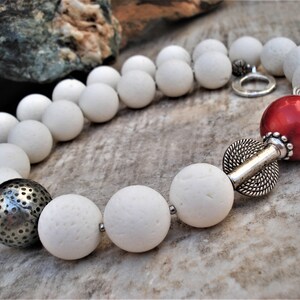 May include: A white stone bead necklace with a red coral bead and silver accents. The necklace is made of natural materials and has a rustic, bohemian style.