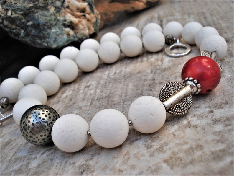 May include: A white stone bead necklace with a red coral bead and silver accents. The necklace is made of natural materials and has a rustic, bohemian style.