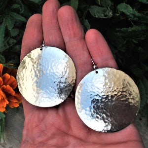 Puede incluir: Un par de pendientes circulares grandes de metal plateado con una textura martillada y con hoyuelos en toda la superficie. Cada pendiente est&aacute; suspendido de un gancho sencillo, creando un acabado met&aacute;lico y reflectante.