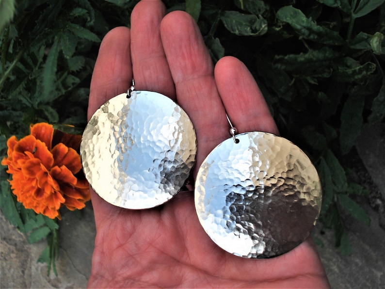 Puede incluir: Un par de pendientes circulares grandes de metal plateado con una textura martillada y con hoyuelos en toda la superficie. Cada pendiente est&aacute; suspendido de un gancho sencillo, creando un acabado met&aacute;lico y reflectante.