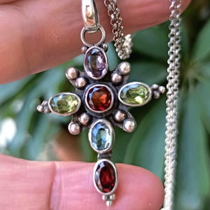 Op de afbeelding: Een zilveren kruis hanger met een verscheidenheid aan edelstenen, waaronder groen, blauw, paars en rood. De hanger is bevestigd aan een zilveren ketting.
