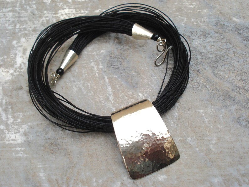 May include: A black leather cord necklace with a large, hammered silver pendant. The pendant is rectangular and has a textured surface.