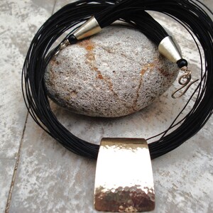 May include: A black leather cord necklace with a hammered gold pendant. The necklace is layered and has a tassel at the end.