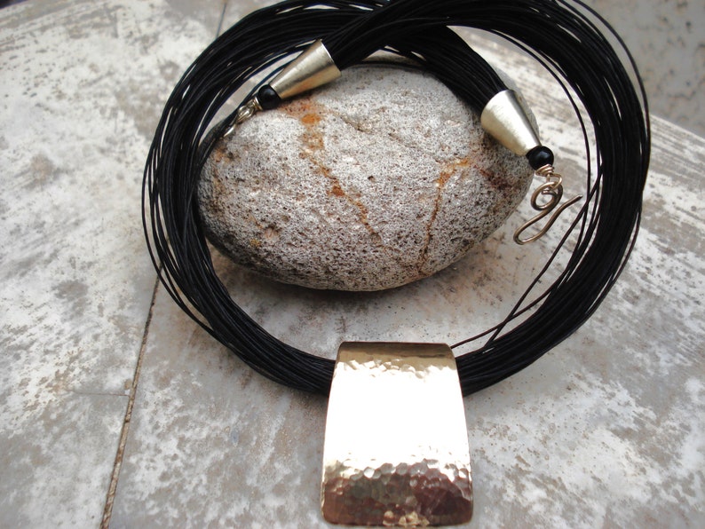 May include: A black leather cord necklace with a hammered gold pendant. The necklace is layered and has a tassel at the end.