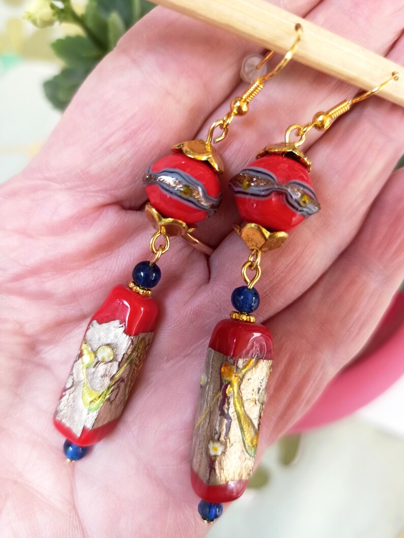 May include: A pair of dangle earrings with red and gold glass beads and blue accents. The beads are decorated with gold leaf and a floral pattern.