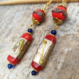 May include: A pair of long, red and gold earrings with blue beads. The earrings feature a red and gold glass bead with a silver and gold design, followed by a long, rectangular red and gold glass bead with a silver and gold design.
