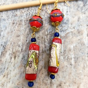 May include: A pair of long, red and gold earrings with blue glass beads. The earrings feature a round red bead with gold and silver accents, followed by a rectangular red bead with gold and silver accents, and a blue glass bead.