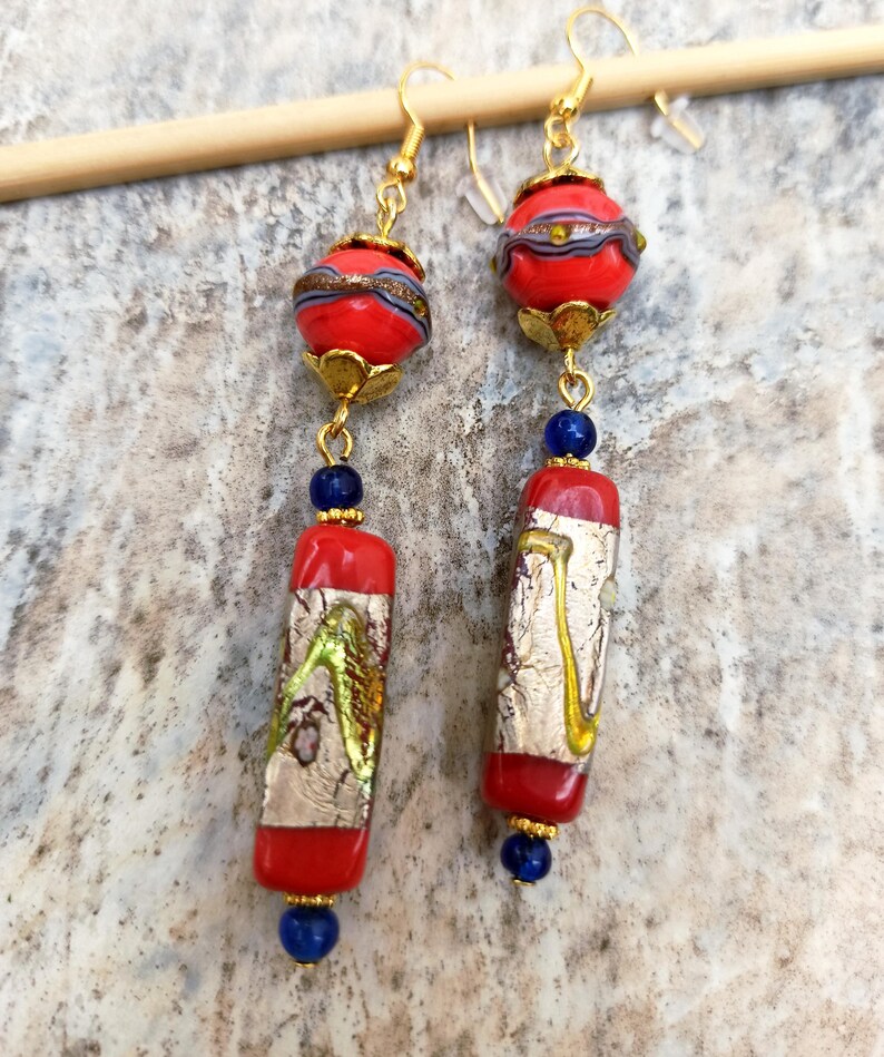 May include: A pair of long, red and gold earrings with blue glass beads. The earrings feature a round red bead with gold and silver accents, followed by a rectangular red bead with gold and silver accents, and a blue glass bead.