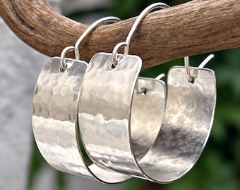 Puede incluir: Un par de pendientes de aro de plata con una banda ancha y curva con un acabado martillado y texturizado. Los pendientes están diseñados con un cierre de gancho simple, mostrando una superficie metálica reflectante.