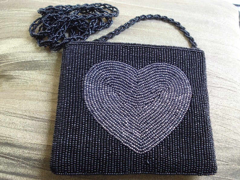 Puede incluir: Un bolso de noche negro de cuentas con un dise&ntilde;o de coraz&oacute;n plateado. El bolso tiene una correa de cadena.