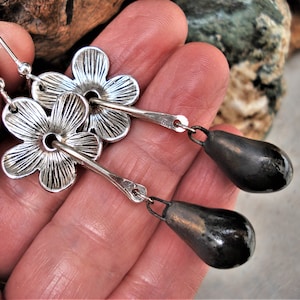 Pode incluir: Brincos de prata com forma de flor e pingentes de lágrima pretos. Os brincos de flor têm uma superfície texturizada e um pequeno furo no centro. Os pingentes de lágrima são lisos e brilhantes.