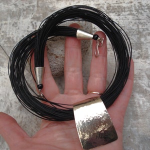 May include: A black cord necklace with a hammered silver pendant. The necklace is made of many thin cords that are tied together at the ends. The pendant is a rectangular shape with a textured surface.