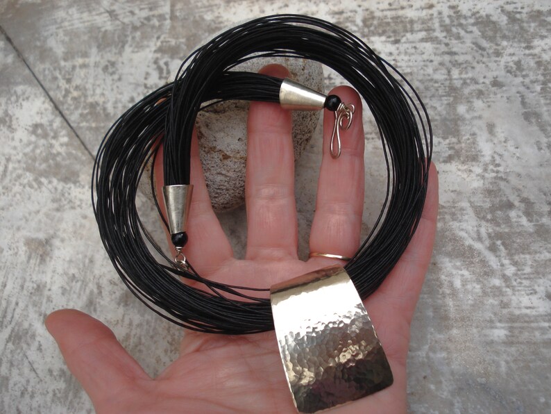 May include: A black cord necklace with a hammered silver pendant. The necklace is made of many thin cords that are tied together at the ends. The pendant is a rectangular shape with a textured surface.