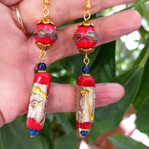May include: Red and gold glass bead earrings with blue accents. The earrings feature a round bead with gold accents and a rectangular bead with gold leaf accents.