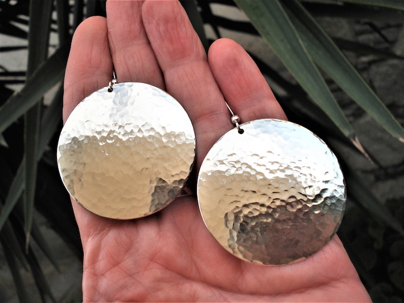 Puede incluir: Un par de pendientes circulares grandes de tono plateado con una textura de superficie martillada y reflectante. Cada pendiente est&aacute; suspendido de un peque&ntilde;o poste de bola de metal en la parte superior, creando una apariencia met&aacute;lica, brillante y distintiva.