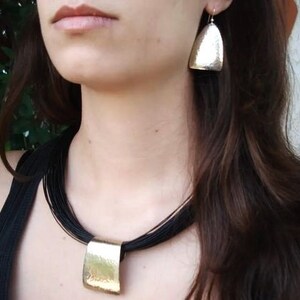 May include: A woman wearing a black choker necklace with a large, rectangular pendant. The pendant is made of hammered metal and has a gold finish. The woman is also wearing silver earrings with a hammered metal design.